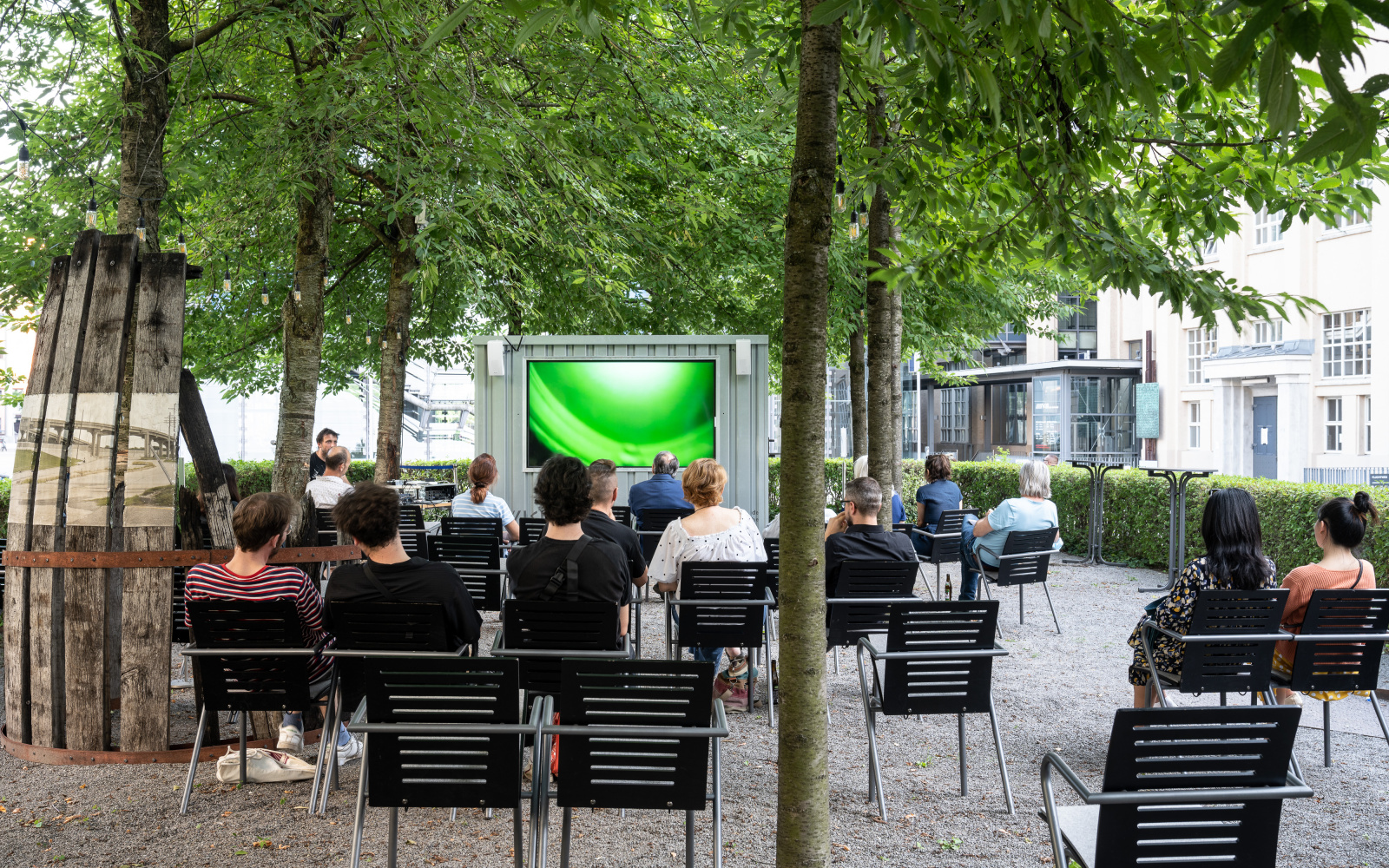 A large screen with people in chairs in front of it outside under green trees.