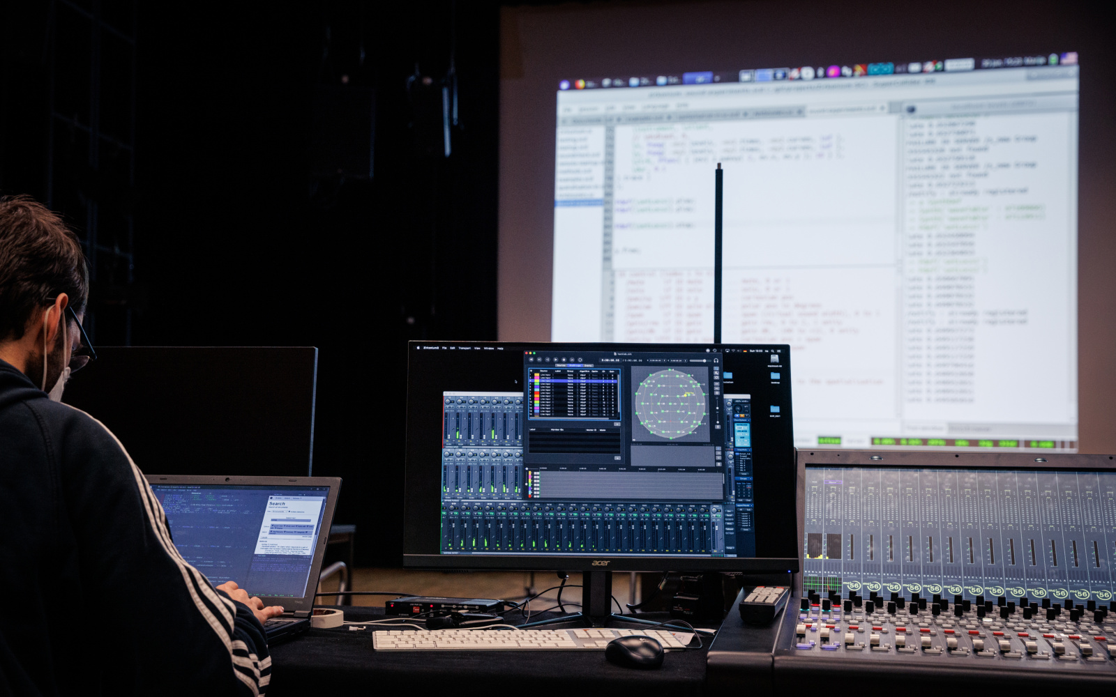 Live Coding Performance: A person is seen from behind, in front of him is a computer screen and a large screen on which a programming code can be seen.