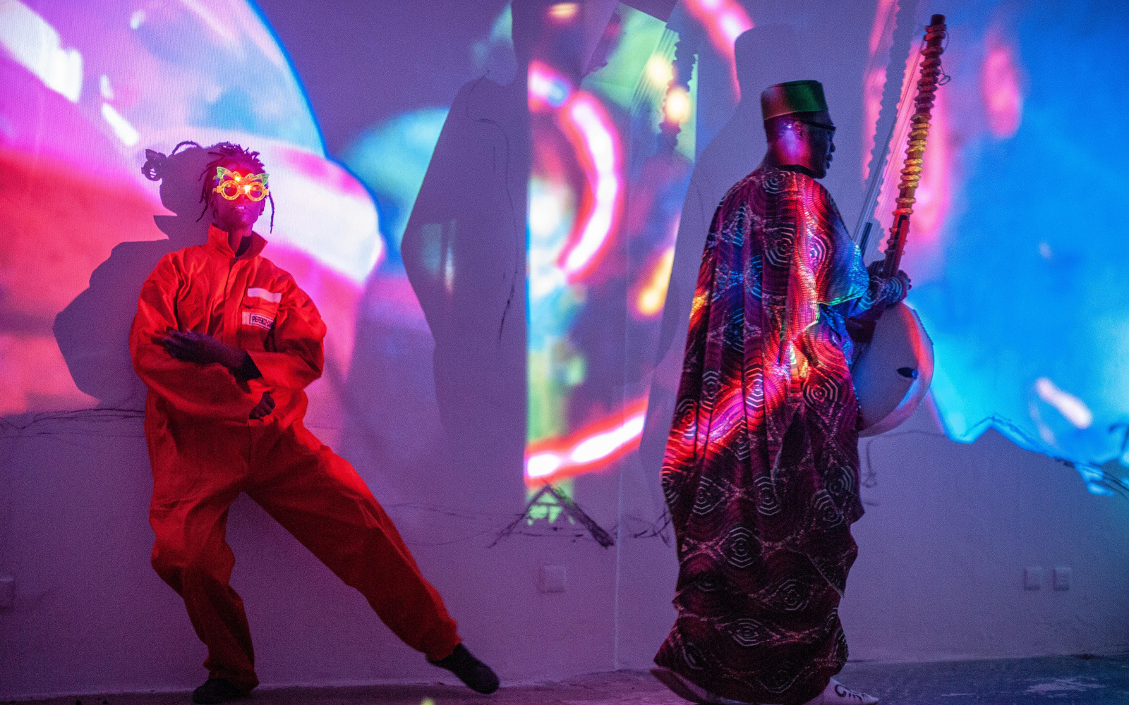 You can see two people illuminated by colorful projections. The left person wears fancy glasses and an orange jumpsuit, the right person wears a traditional robe and has an instrument in his hand.