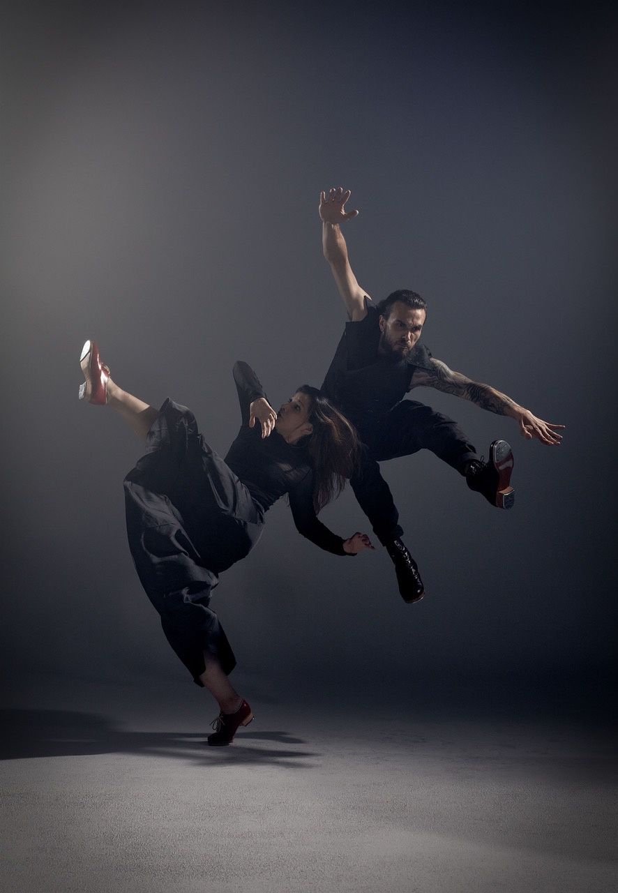Zu sehen sind zwei Tänzer:innen in schwarzer Kleidung vor dunkelgrauem Hintergrund