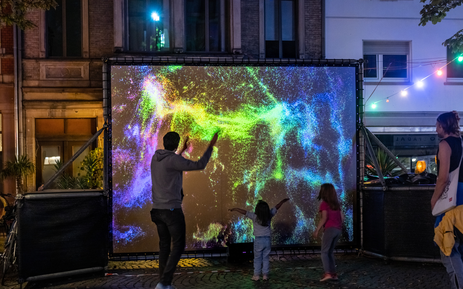 In the pedestrian zone there is a screen with brightly colored particles, and adults and children move in front of it.