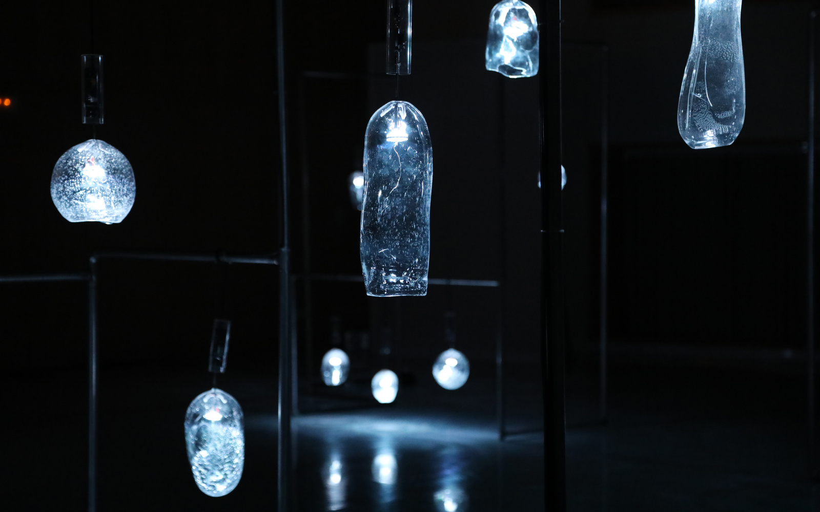 Verschiedenförmige Glasgefäße hängen in einem dunklen Raum und leuchten schwach.