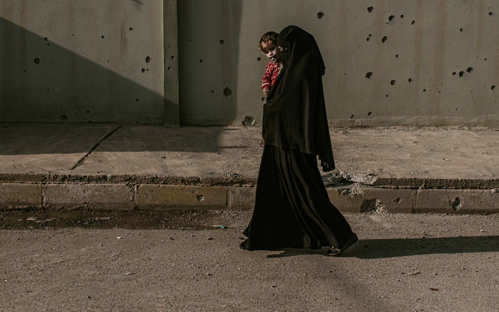 Eine in schwarz verhüllte Frau mit Kind läuft vor einer von Feuerwaffen durchlöcherte Wand.