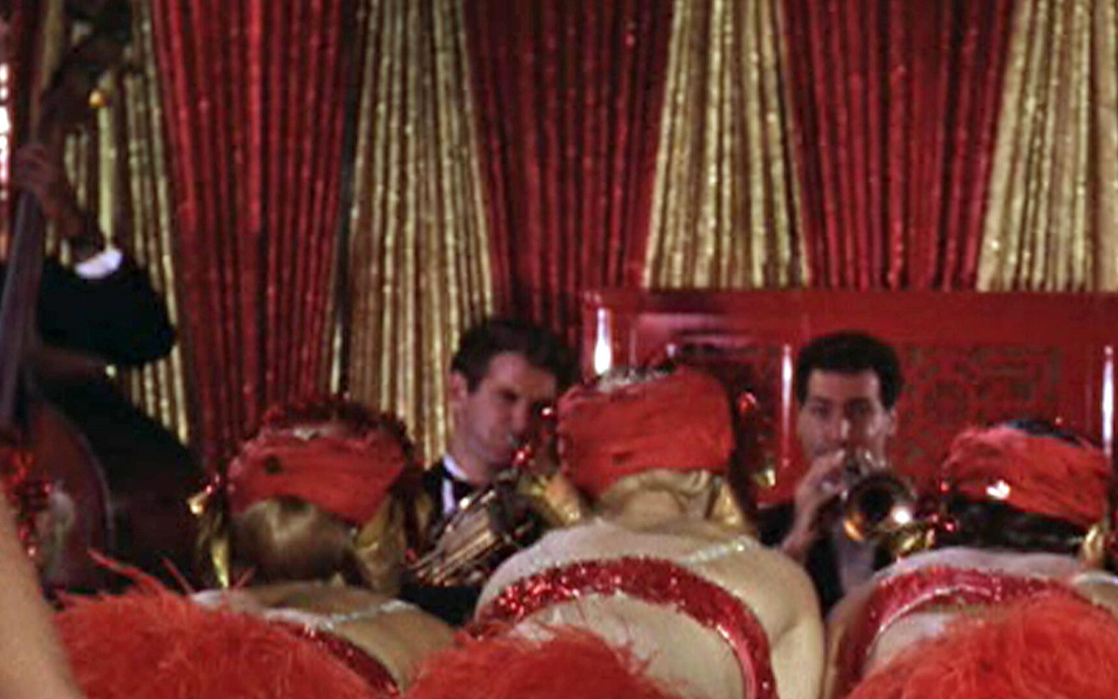 Two trumpet players are seen in front of a golden and red curtain. In the audience sit three women with red headdresses