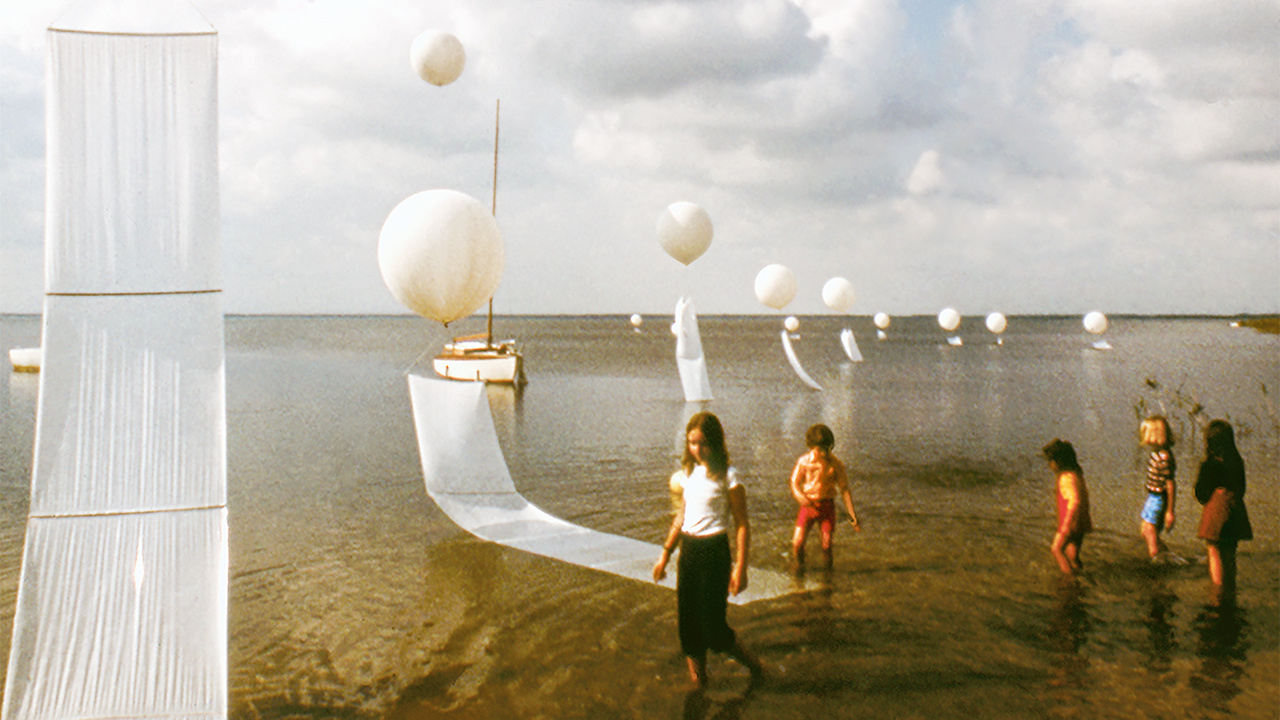 Zu sehen sind fünf Kinder, die durch Wasser laufen. Auf der linken Seite sind weiße Luftballons zu sehen.