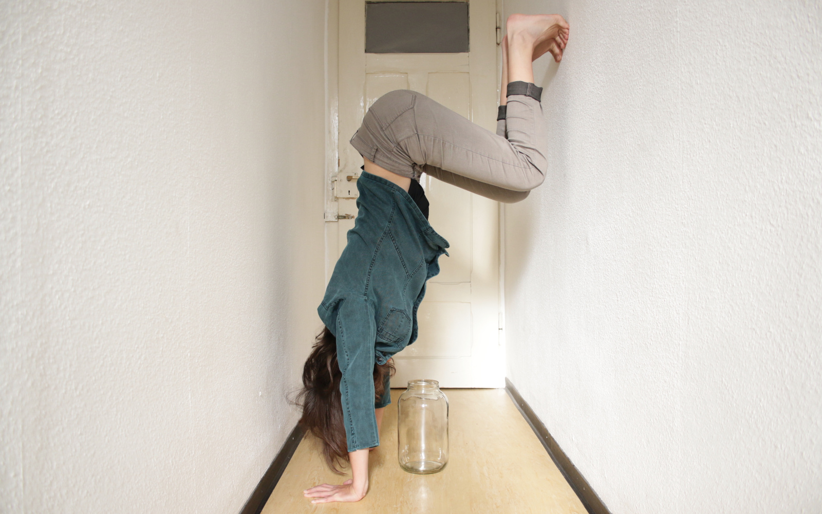 A person is doing a handstand against the wall in a small hallway, under her is an empty jar.
