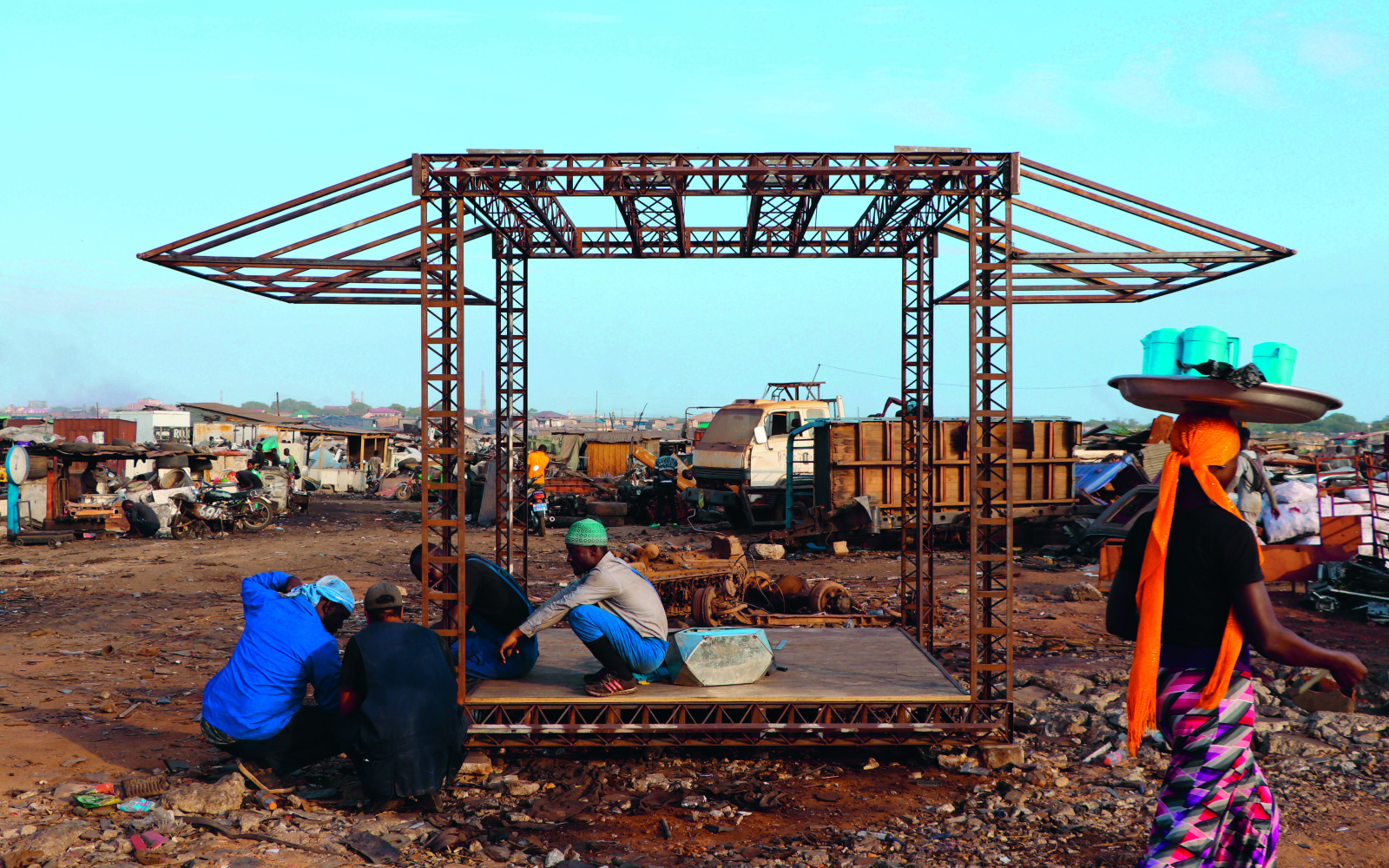 Something is being built in a scrap yard: A walk-in square made of metal struts. A builder is squatting in it.