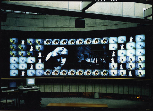 The photo shows a large screen that is composed of many smaller screens and thus shows a large image. That of a young woman looking into the camera