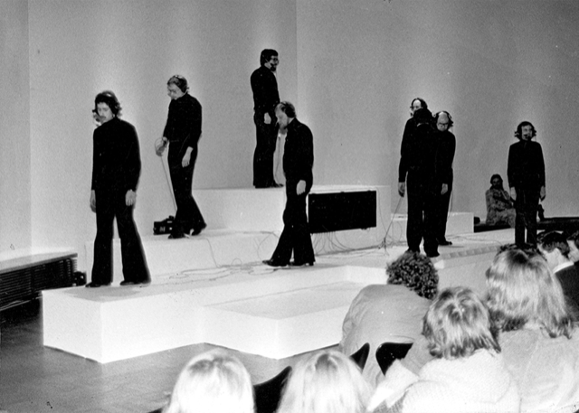 »Homo lusus – der gespielte Mensch« von Walter Giers. Es sind neun in schwarz bekleidete Personen zu sehen, die auf Podesten stehen. 
