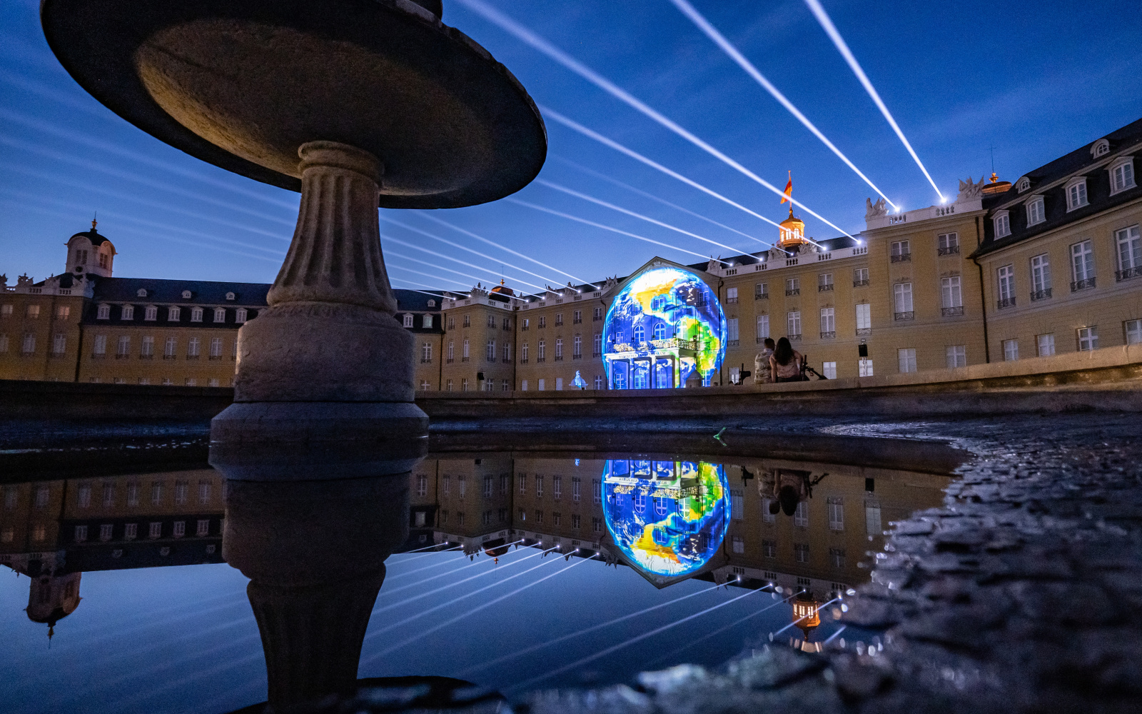 Zu sehen ist die beleuchtete Fassade des Karlsruher Barockschlosses, in deren Mitte sich eine Weltkugel befindet.