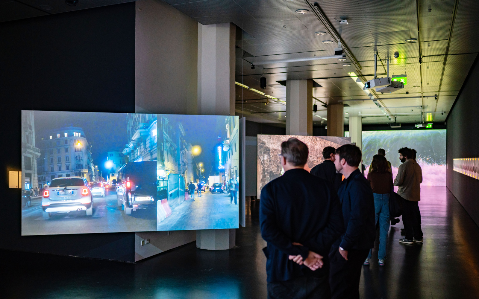 Zu sehen sind zwei Personen vor einem Bildschirm, auf dem ein Auto auf der Straße gezeigt wird.Ausstellungsansicht »Marijke van Warmerdam. Then, now, and then«.