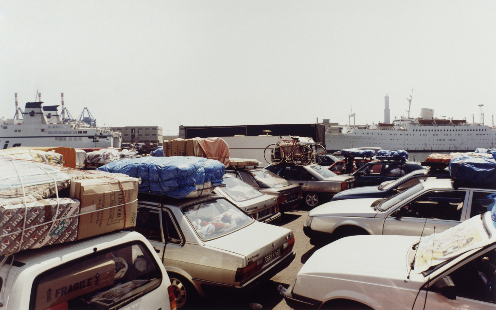 Photography of several parked cars, all fully loaded. In the background are two large cruise ships.