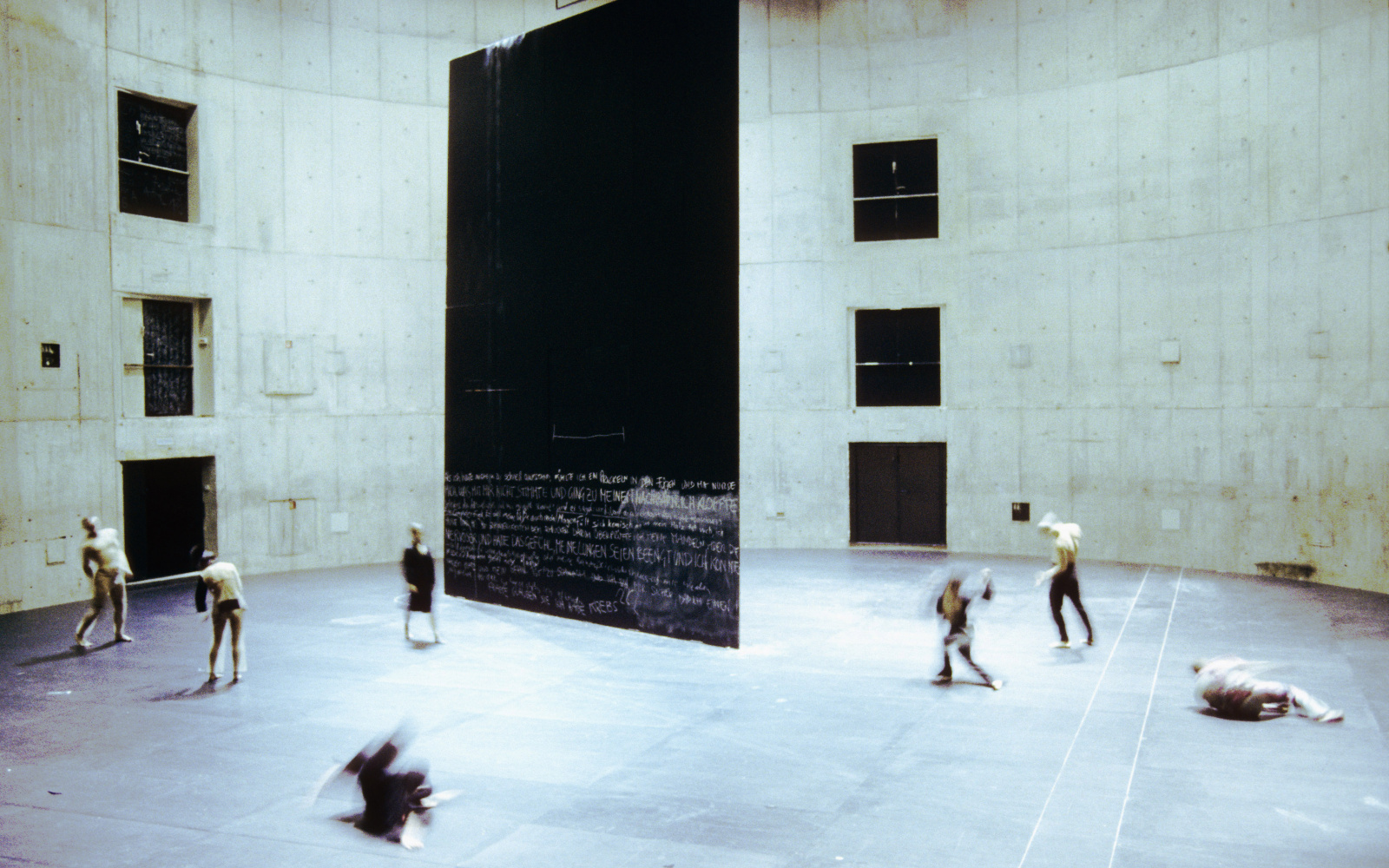 Seven dancers move around a large rectangular black wall that is structured a theatrical space.