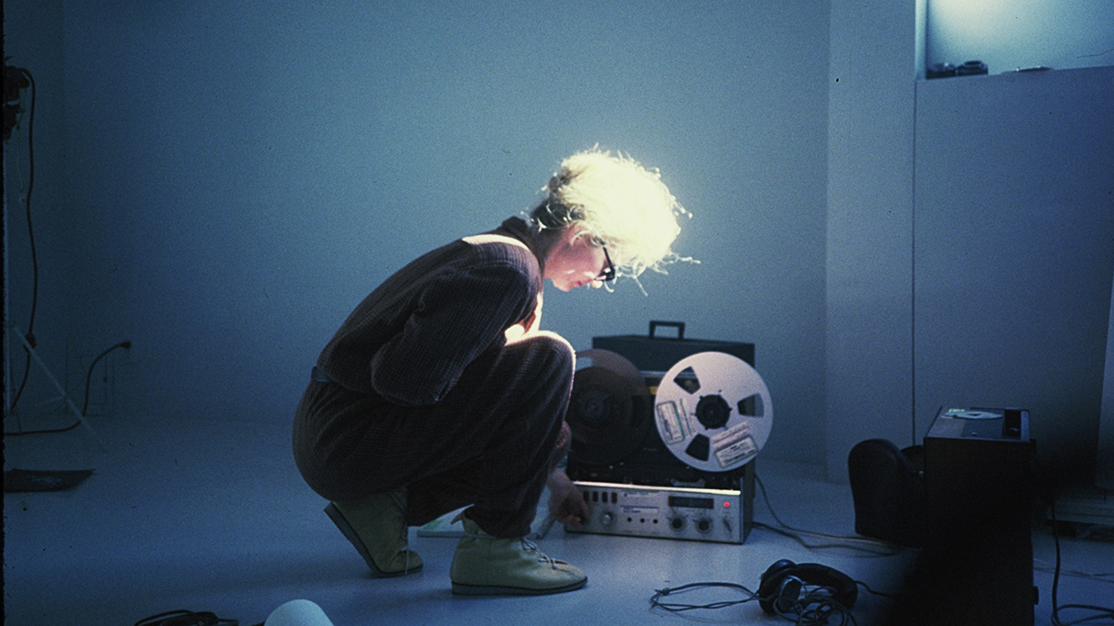 In the picture, a woman is sitting in a crouching position and adjusting something on a receiver that is standing diagonally next to her. Around her are some technical objects on the floor and she herself has sunglasses on and is illuminated through a win