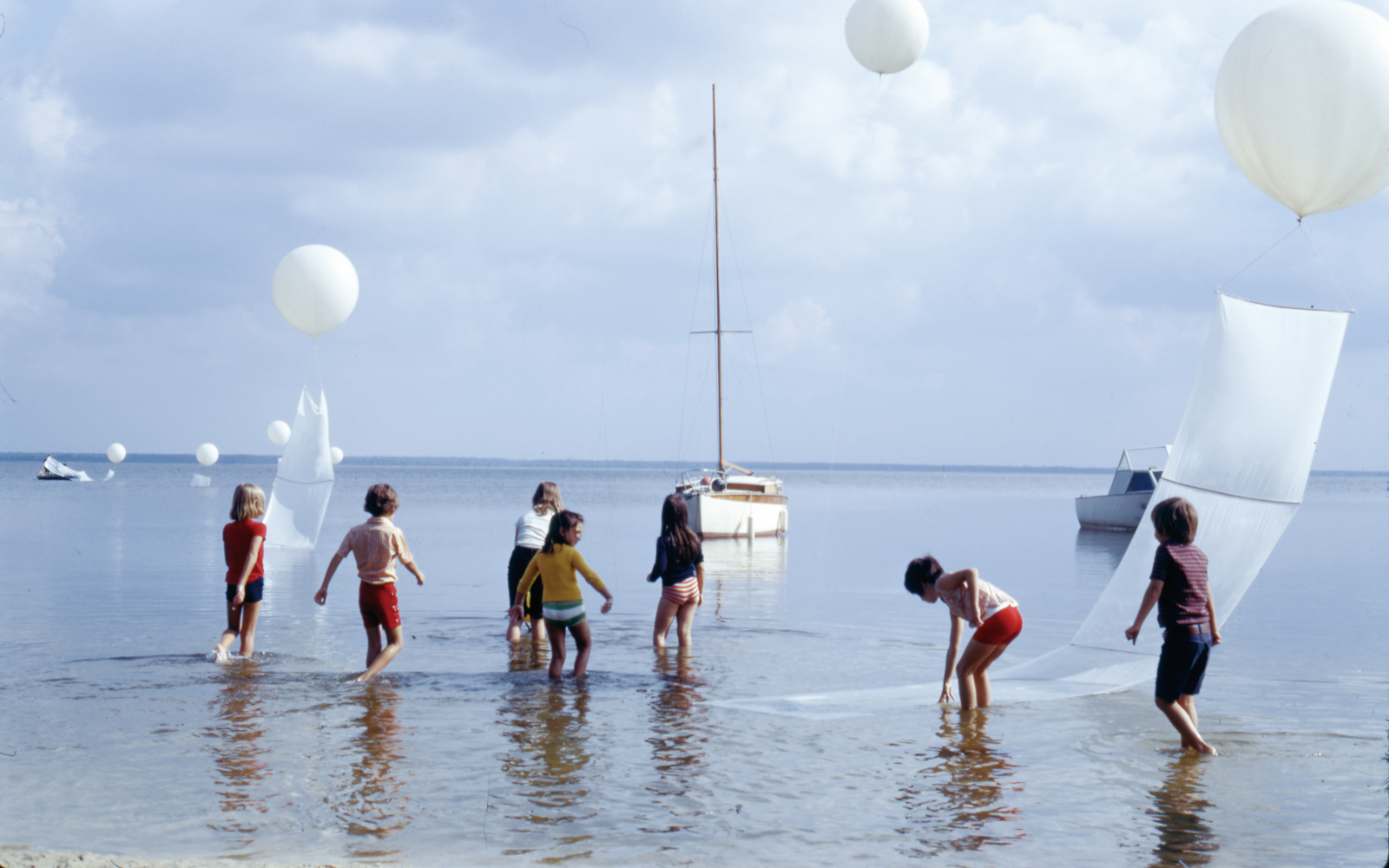 You can see several children running through water. Above them are white balloons. In the background you can see boats. 