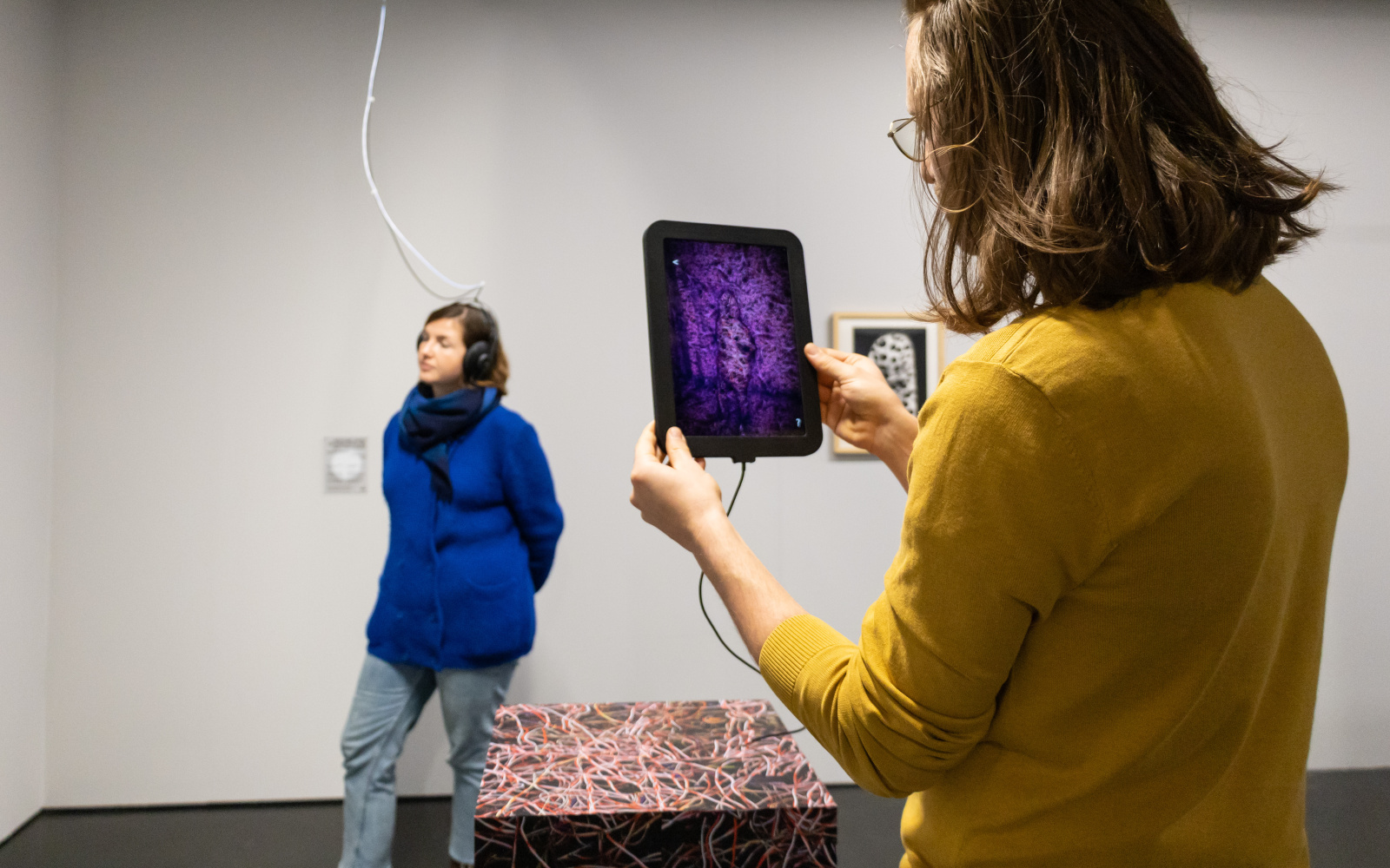 to see are two women. One is standing in the background and is photographed by the one in front through a tablet.