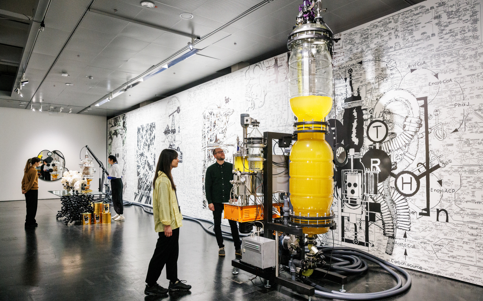 A ceiling-high, elongated glass vessel containing a yellow liquid can be seen. Several cables or hoses lead to smaller glass vessels that also contain liquid. Large hoses are lying on the floor. Two people are looking at this.