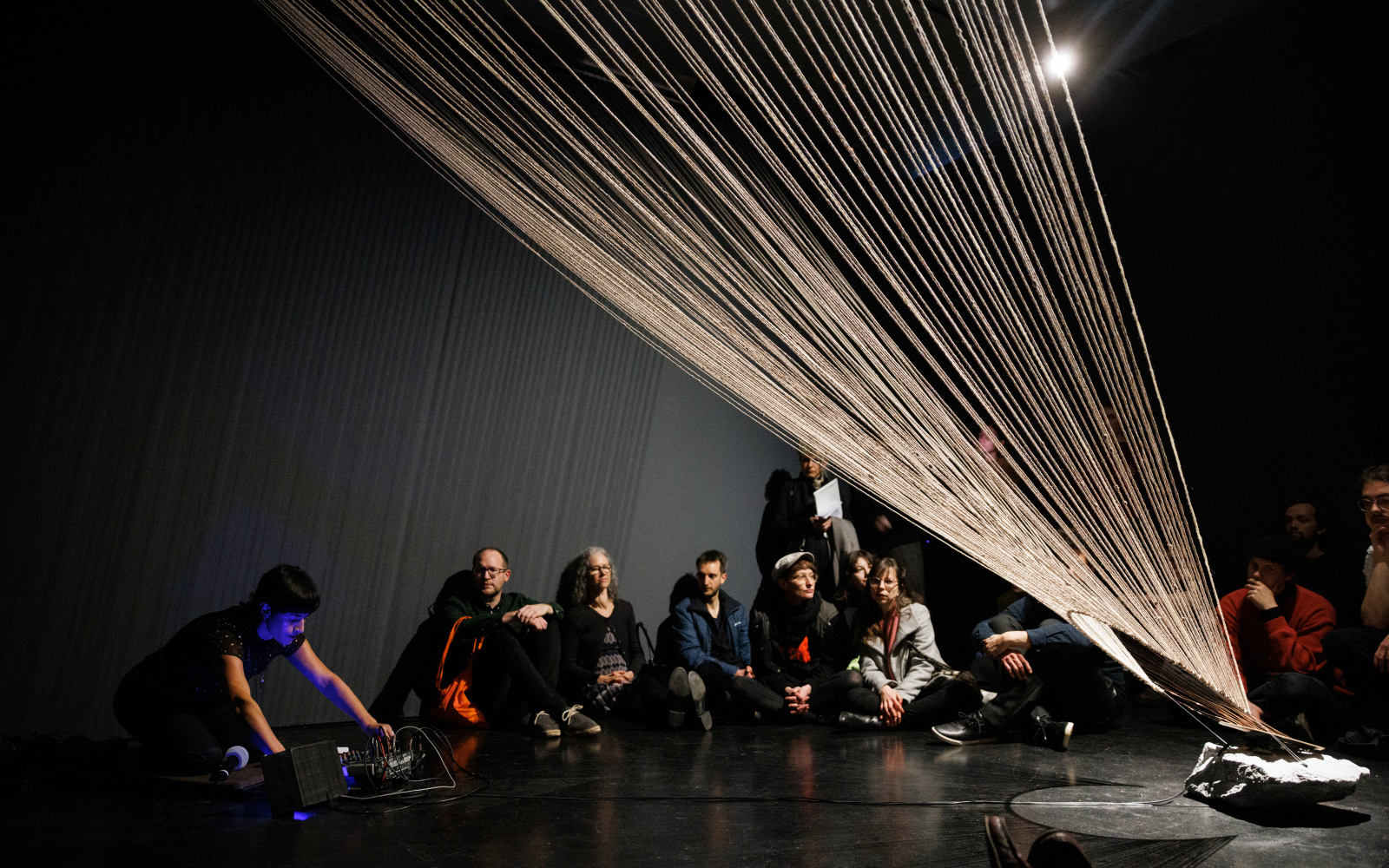 You can see a large, fanned net of threads that reaches up to the ceiling. In the background, several people are sitting against a wall.