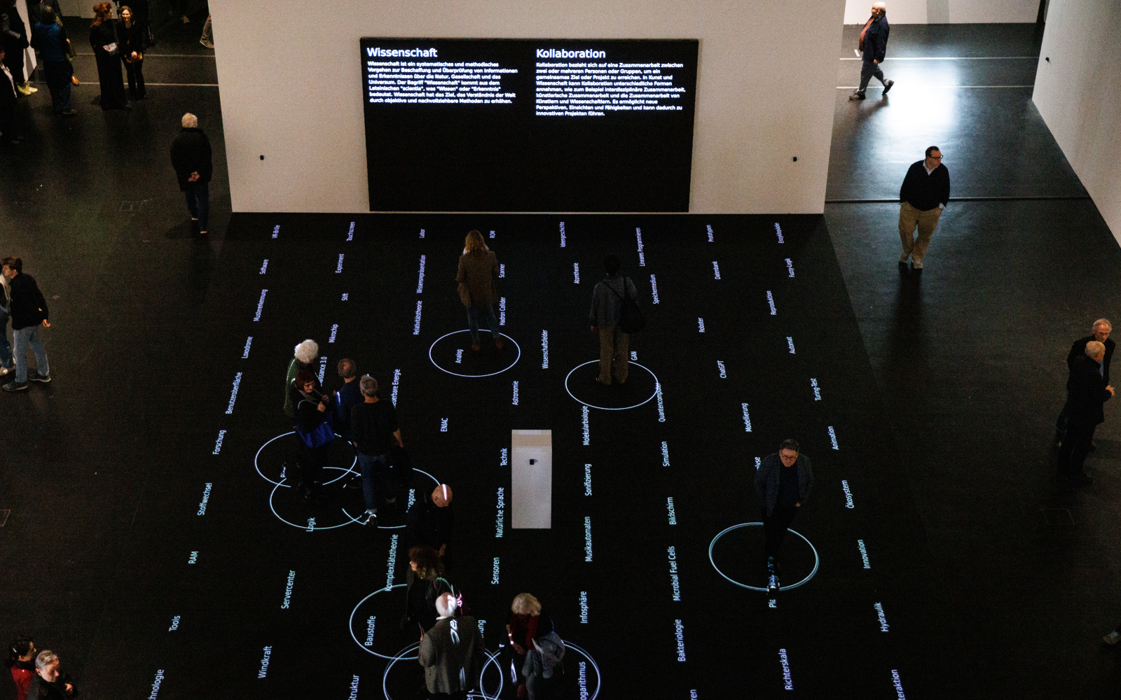 You can see several people on a black surface. On this surface there are words. Around the people are white circles. In the background are two screens.
