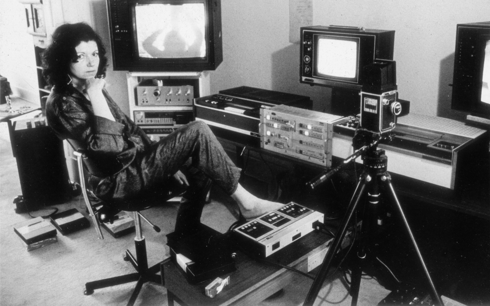 The photo is black and white and shows Ulrike Rosenbach, the media artist, in her studio in the 70s. Ulrike Rosenbach is sitting on a swivel chair and is surrounded by camera equipment and tube screens. 