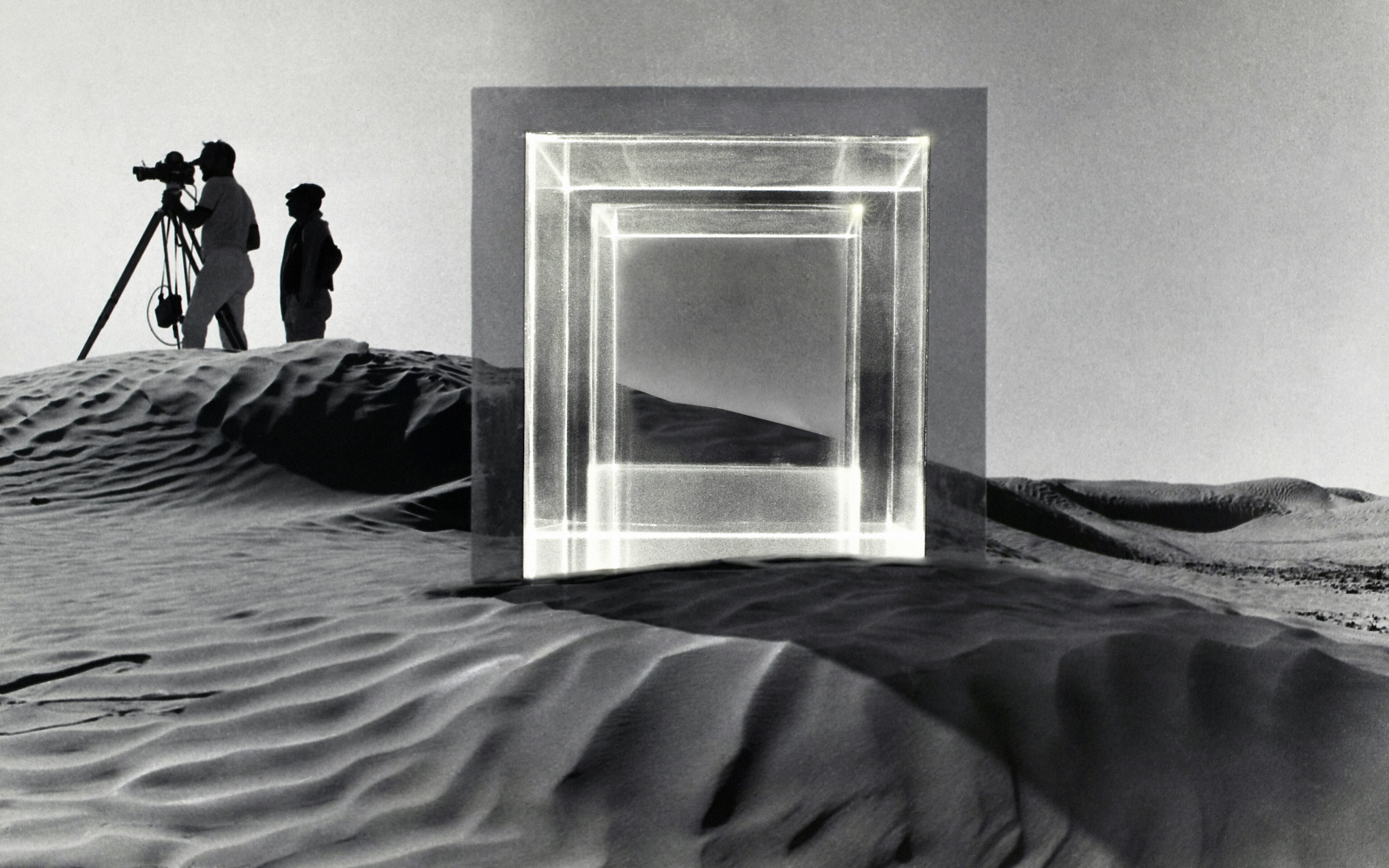 In einer Wüstenlandschaft um 1968 fanden Dreharbeiten zu dem Film »Tele-Mack« statt. Im Vordergrund steht eine große quadratische Glasskulptur und im Hintergrund stehen zwei Männer mit einer Filmkamera. Das Bild ist schwarz-weiß.
