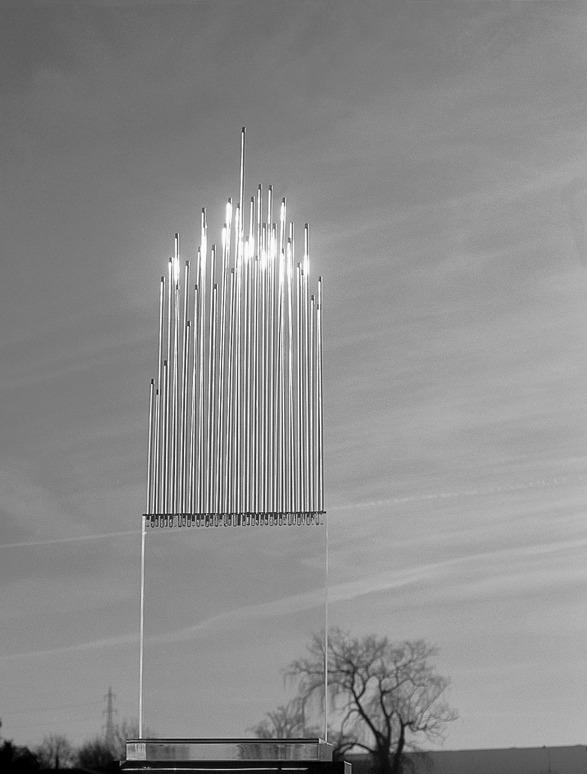 In the middle of a landscape stands a large glass sculpture. The lower part of the sculpture is made of transparent glass and the upper part is made of glass, which looks like a harp and has no clear edge upwards. The image is black and white.