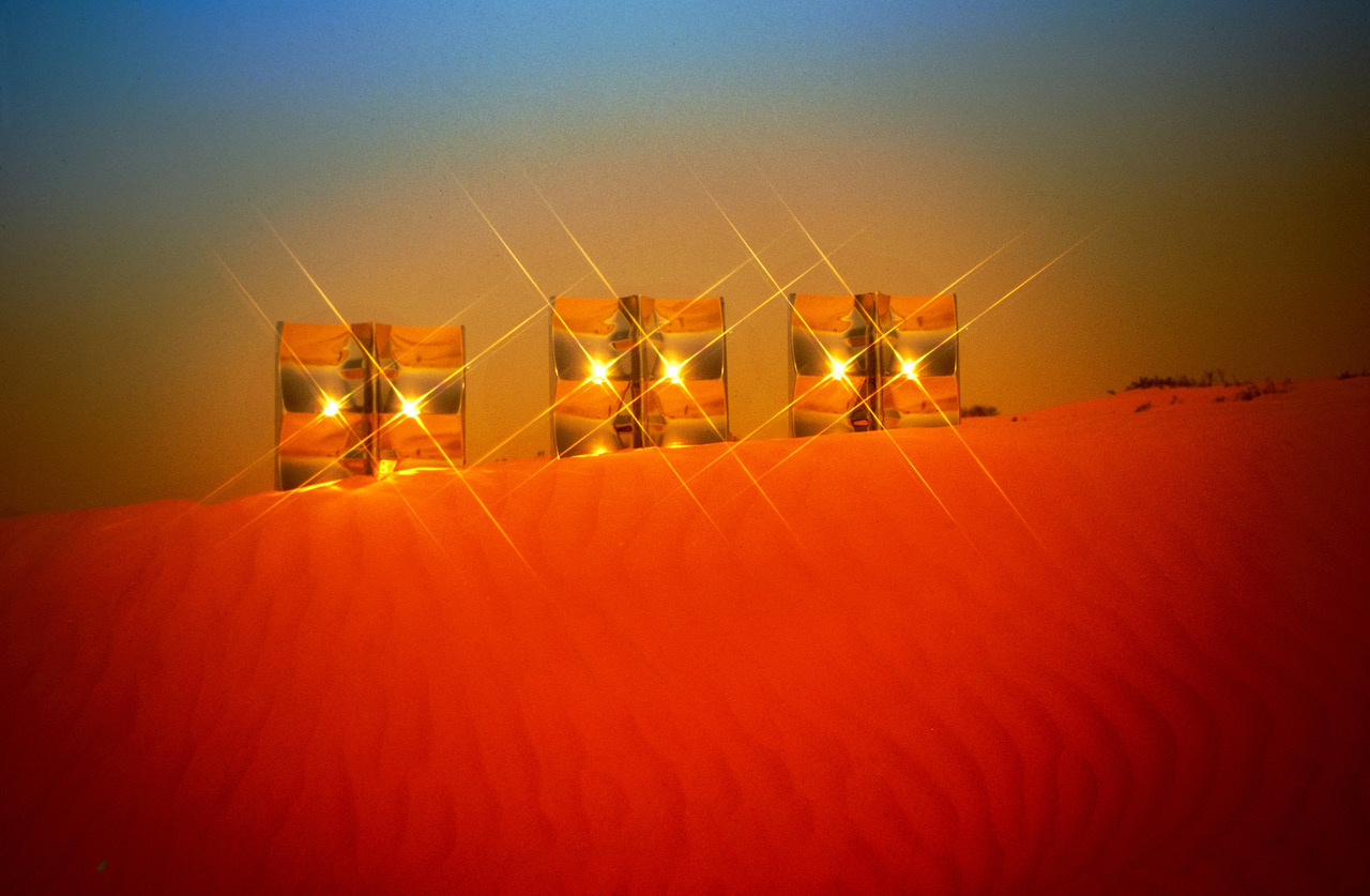 Zu sehen ist eine rote Wüste und blauer Himmel. In mitten des Bildes und auf einer Düne stehen drei abstrakte Quadrate, die wie Spiegel, das Licht reflektieren.