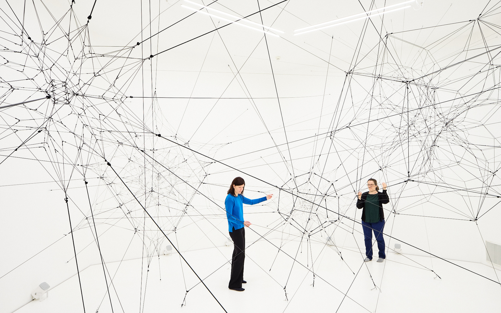 »Algo-r(h)i(y)thms« von Tomás Saraceno. Zu sehen ist ein großes Netz aus Fäden in einem weißen Raum. Zwei Personen stehen in diesem Raum und berühren diese Fäden.