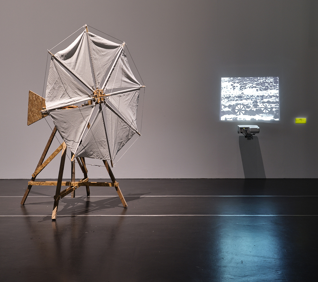 »Windmill 03 (for Walter Segal)« von James Bridle. Zu sehen ist eine Windmühle aus Holz, bespannt mit weißen Tüchern. Auf der rechten Seite ist eine kleine schwarz-weiß Projektion zu sehen.