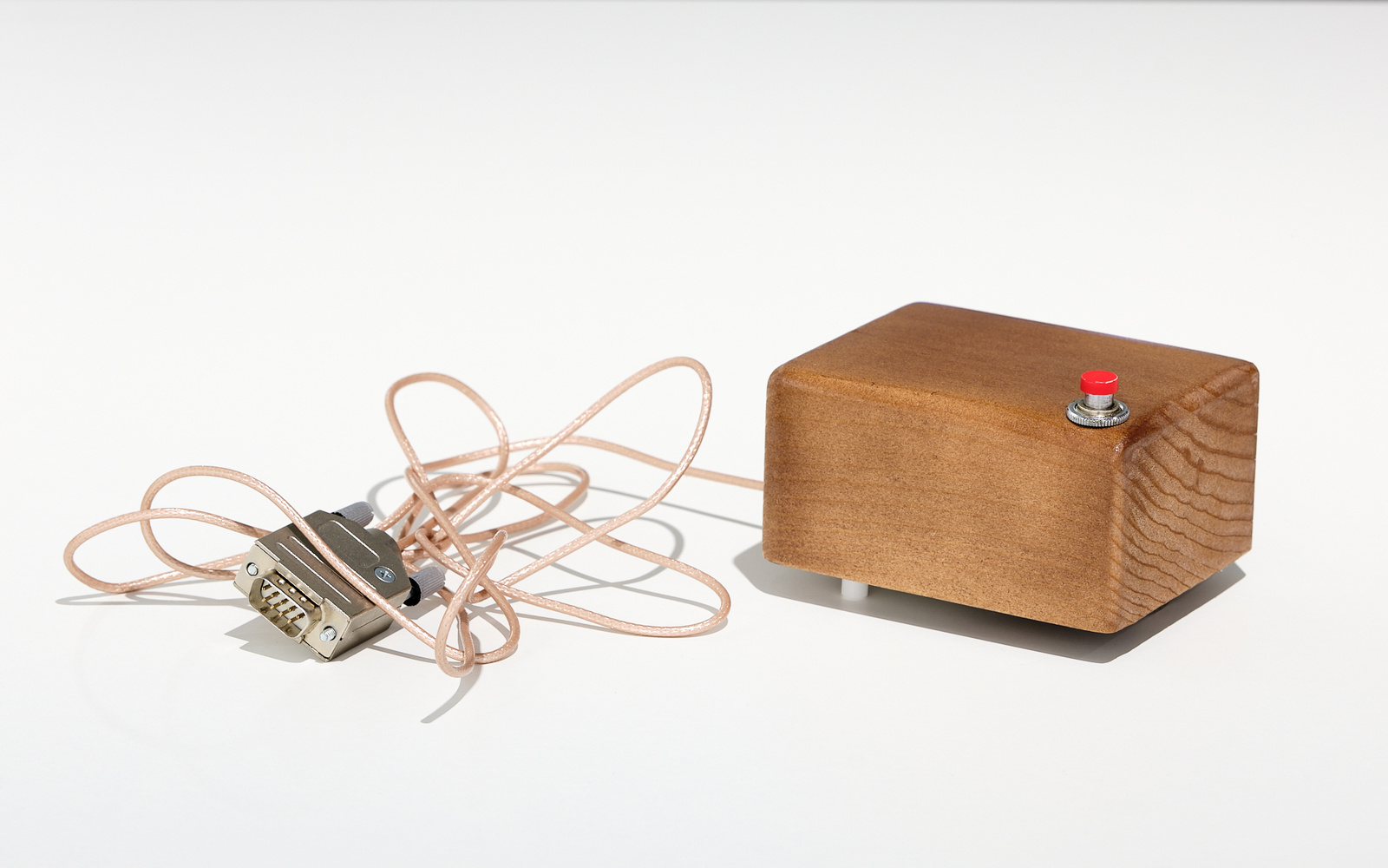 »Mouse« von Douglas C. Engelbart. Zu sehen ist ein Holzblock. Vorne am äußeren Rand befindet sich ein roter Knopf. An dem Holz ist ein Kabel mit Stecker befestigt.
