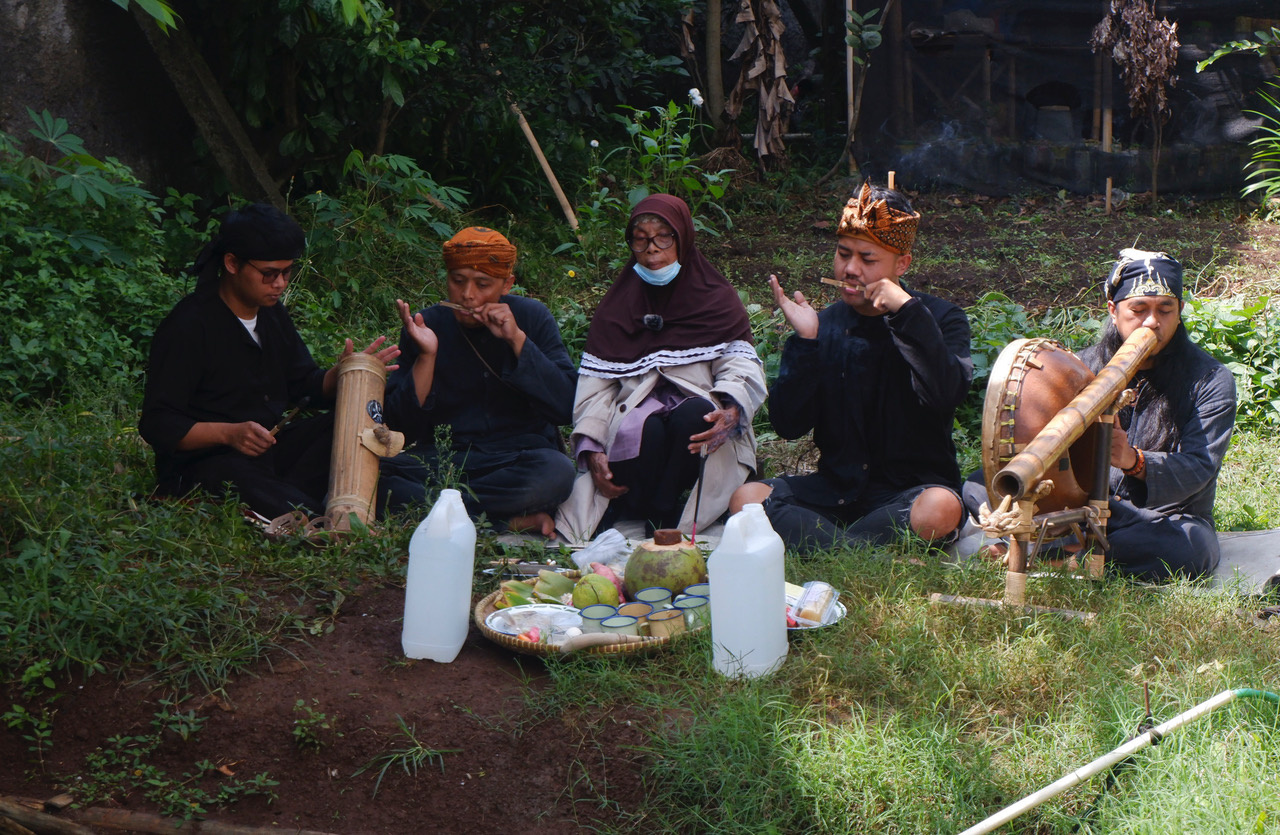 You can see five people sitting on the ground in the forest. In front of them is a tray with cups and coconuts. The people have objects made of wood in their hands and mouths.