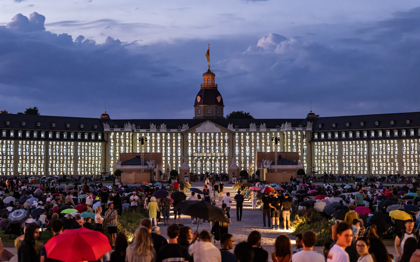 The façade of the baroque palace in Karlsruhe can be seen. Peter Weibel's show "We are data" will be projected. The castle has a dark background and the words "Daten" (data) are written many times in white letters.