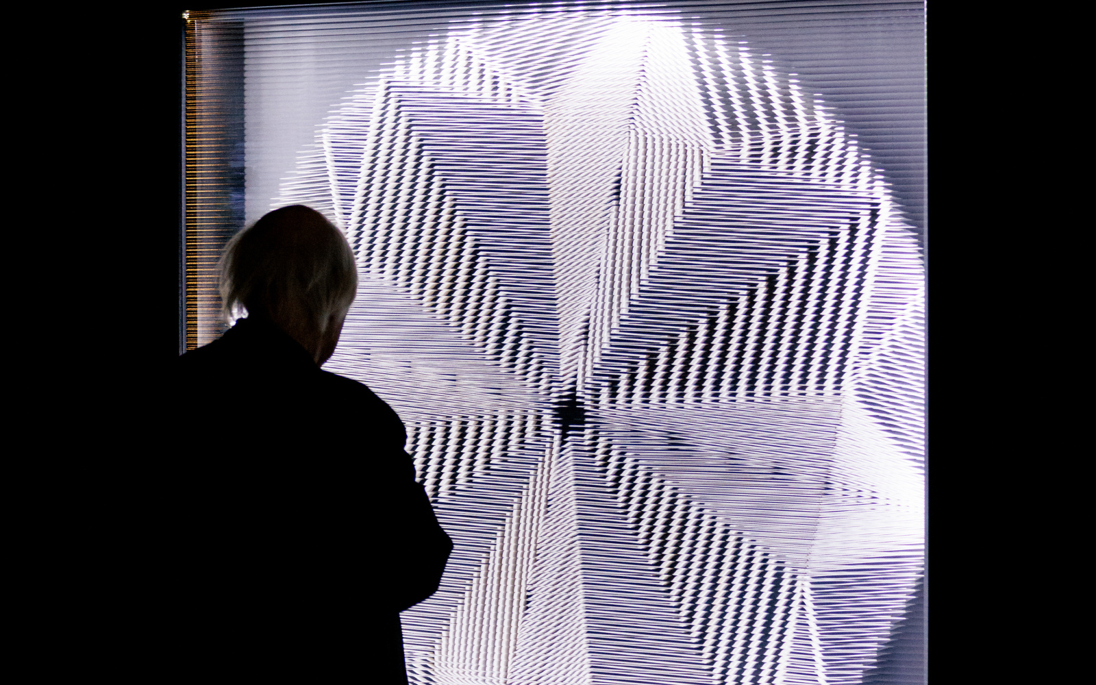 An elderly man stands in front of a round luminous disc, which creates a pattern because the glass in front of it is cut, and looks at it.