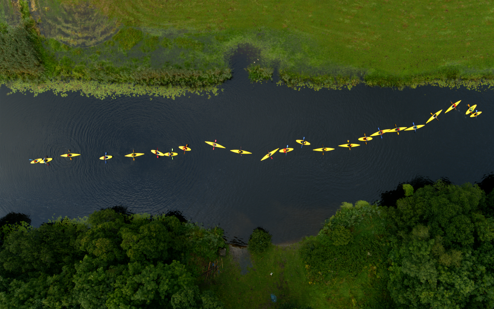 Zu sehen ist eine grüne Landschaft durch die sich ein Fluss schlängelt. Das Bild zeigt aus der Vogelperspektive, wie neun Menschen in gelben Regenjacken und gelben Kanus, den Fluss entlangpaddeln.