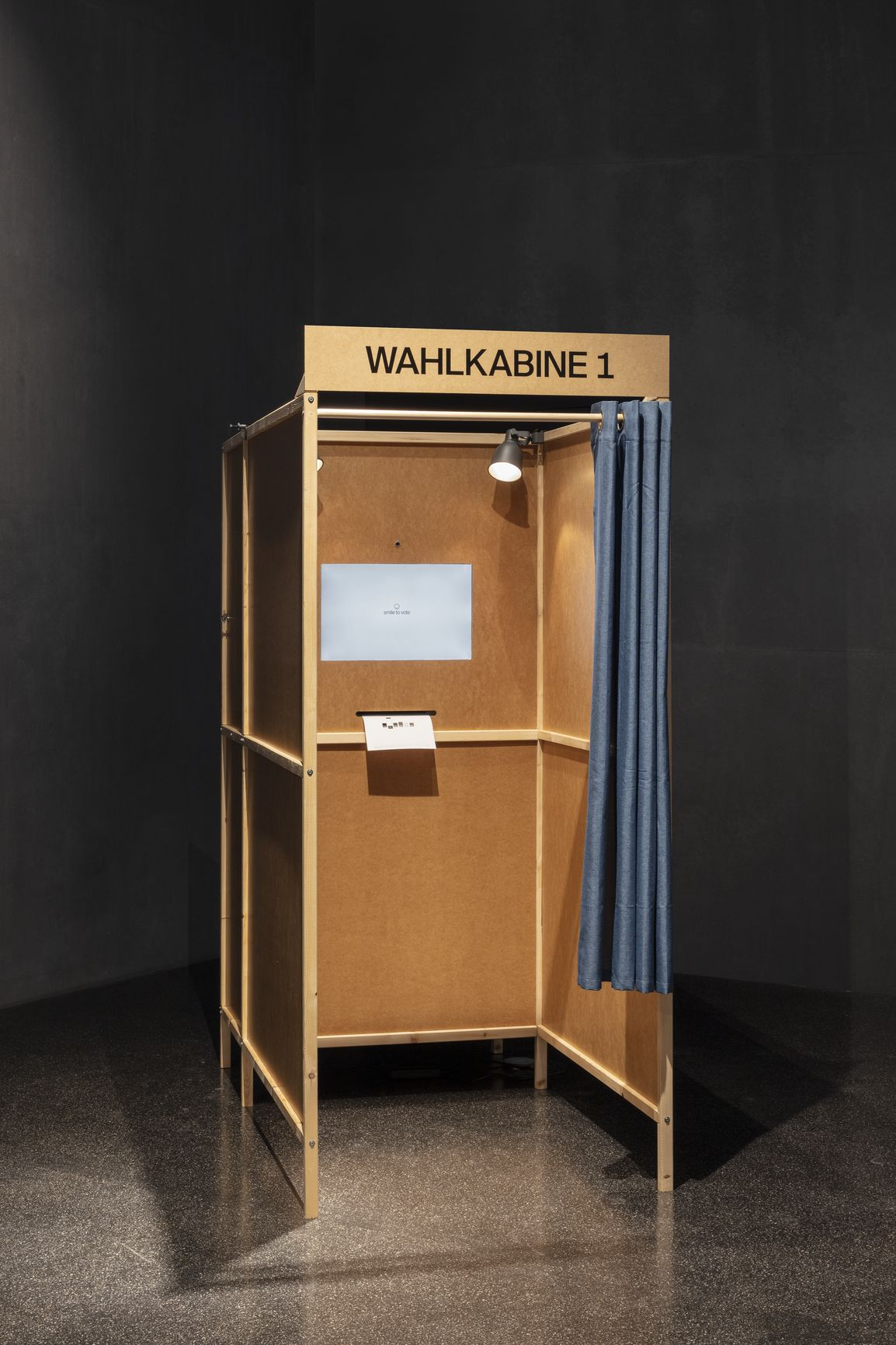 Das Bild zeigt eine Wahlkabine aus Holz mit einem blauem Vorhang. Sie ist Teil des Gesamtkunstwerks »Smile to Vote – Political Physiognomy Analytics«, ein künstlerisch-wissenschaftliches Forschungsprojekt, das die Auswirkungen von KI-basierten biometrisch