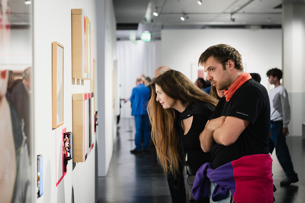 A woman and a man look at pictures on a wall. In the background, several people can be seen walking through the exhibition.