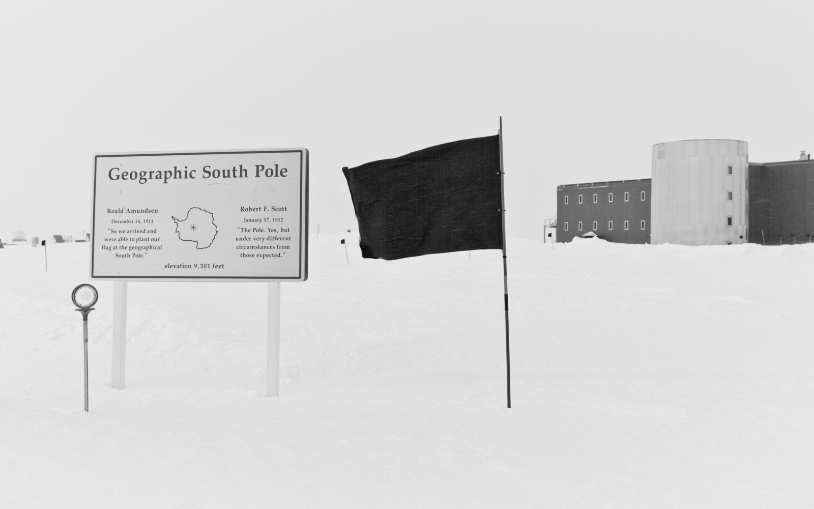 Das Bild zeit eine schwarze Flagge im Schnee neben einer Infotafel über den Südpol und einem Haus, was im Hintergrund zu sehen ist.