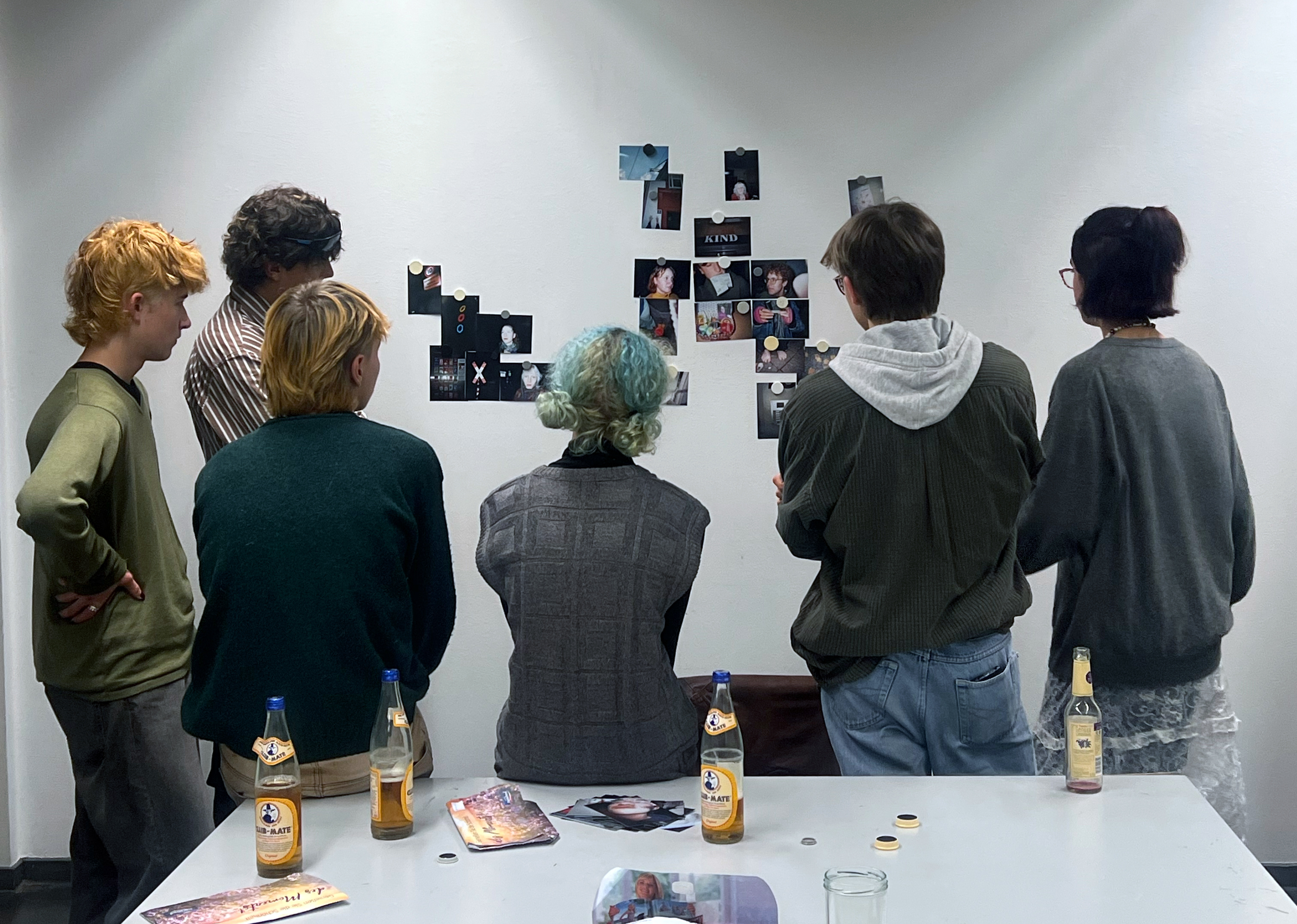 Fünf junge Kunststipendiat:innen sind von hinten zu sehen, sie stehen vor einer weißen Wand an der Fotos hängen