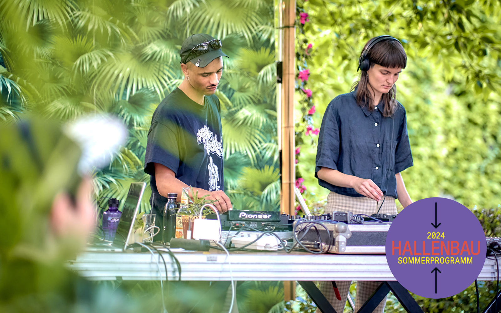 A DJ and a DJane are standing outside at a mixing desk, with lots of greenery in the background.