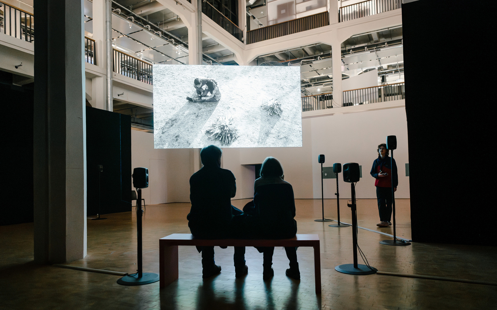 Two people are seated on a bench in front of a screen. 