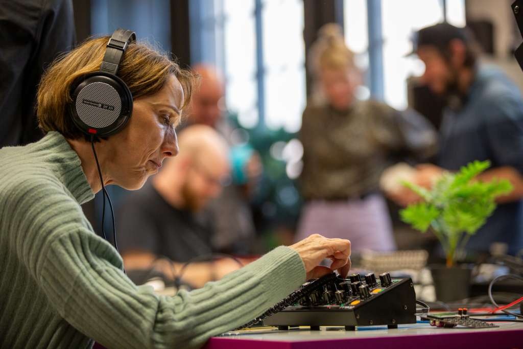 An older person is sitting at a table with headphones on and is turning knobs. More people can be seen in the background, out of focus.