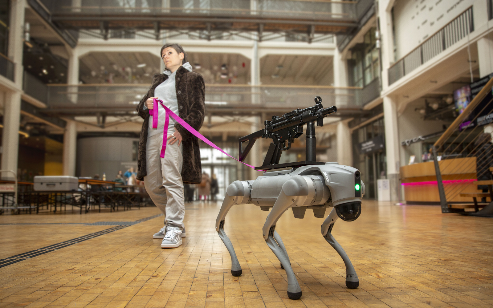Ein Roboter Hund mit einem Sturmgewähr auf dem Rücken, der von einer pinken Leine von einer Person mit braunen Haaren und Pelzmantel gehalten wird.