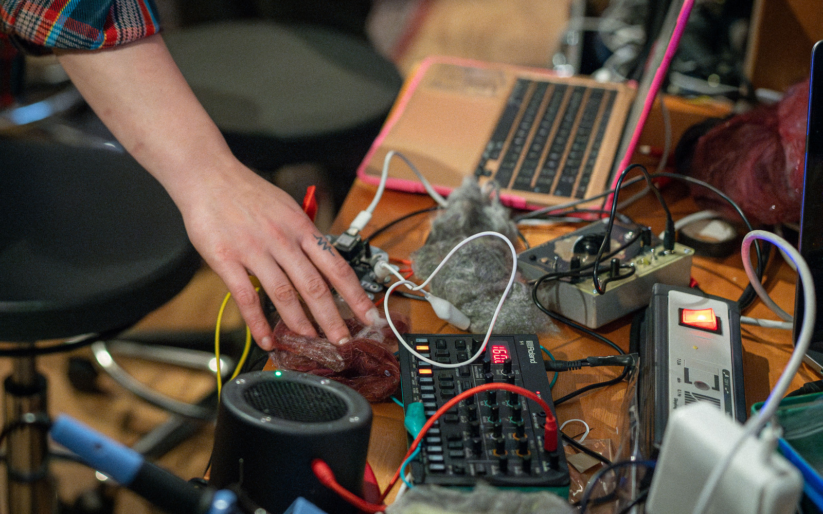Das Bild zeigt eine Hand, die verschiedene elektronische Musikinstrumente und Synthesizer auf einem Tisch mit Kabeln, einem Laptop und weiteren Audiogeräten bedient.