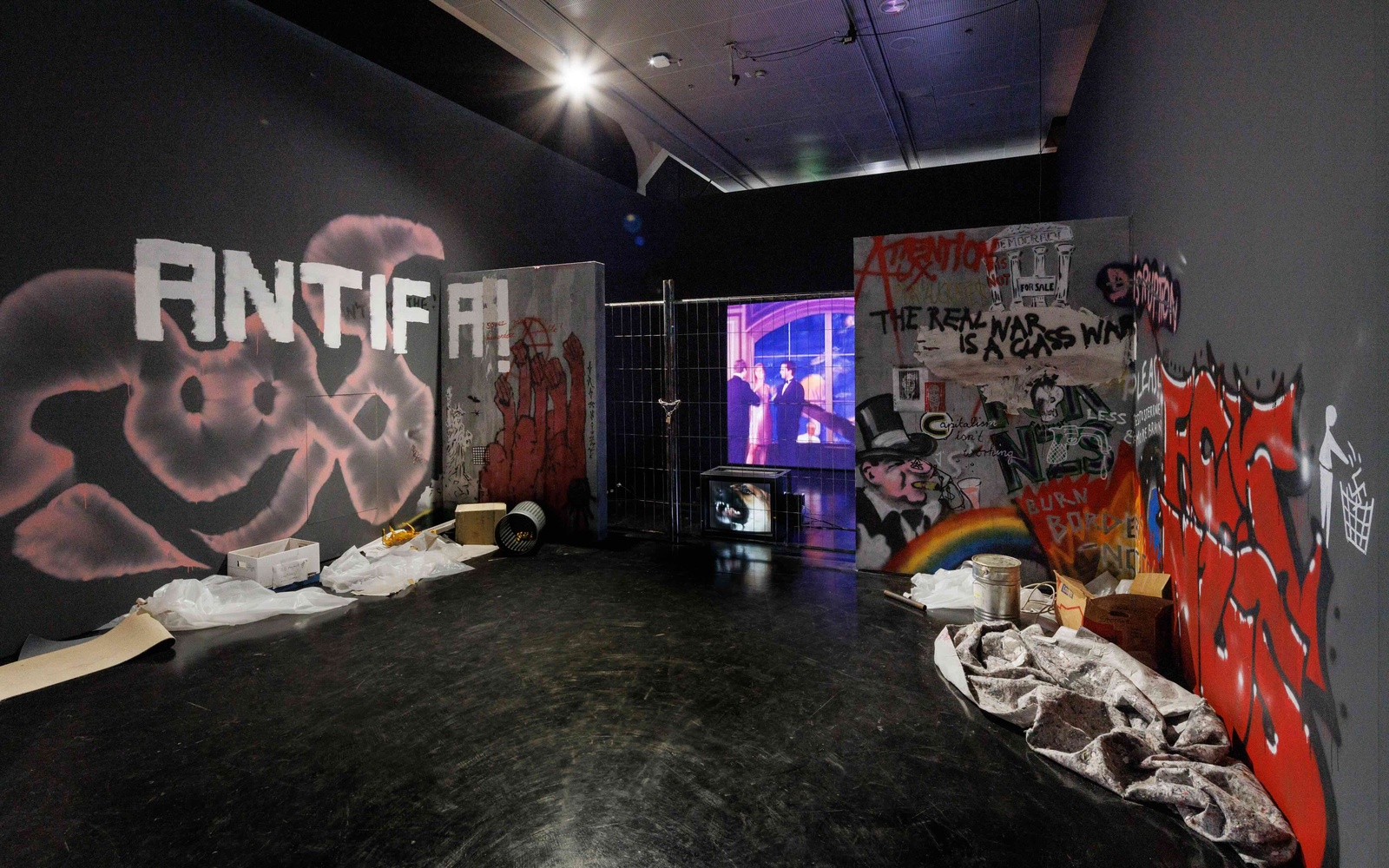 Installation in einem Ausstellungsraum mit politischen Graffiti, Absperrgitter, Projektionsbildschirm und verstreuten Materialien auf dem Boden.