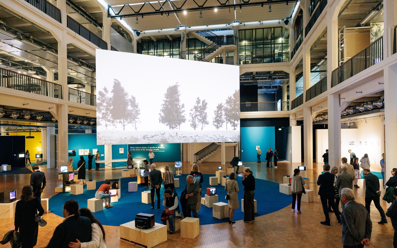 Visitors view a multimedia exhibition featuring vintage TVs and a large-scale video projection in an open museum space.