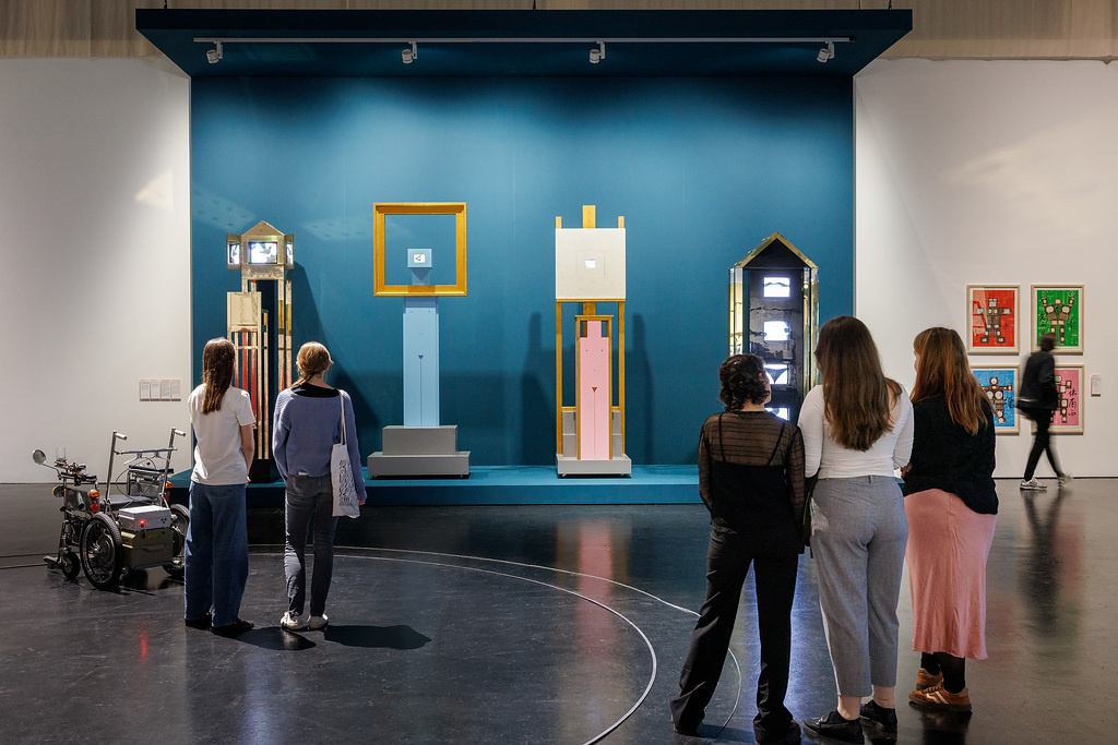 Blick in die Ausstellung The Story That Never Ends, 5 Personen schauen Kunstwerke an, blauer Hintergrund