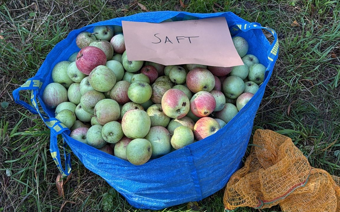 Ein großer Sack Äpfel ist zu sehen, welcher auf einer Wiese steht und mit einem Schild versehen ist, auf dem Saft steht.