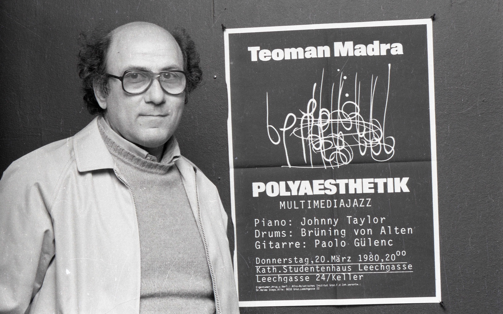 Zu sehen ist eine Person mit Brille, die neben einem Plakat steht. 