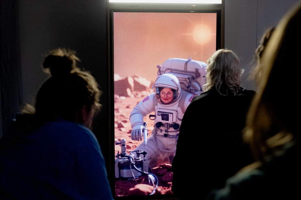 Zu sehen sind mehrere Personen, die vor einem Bildschirm stehen, der eine Astronautin zeigt.
