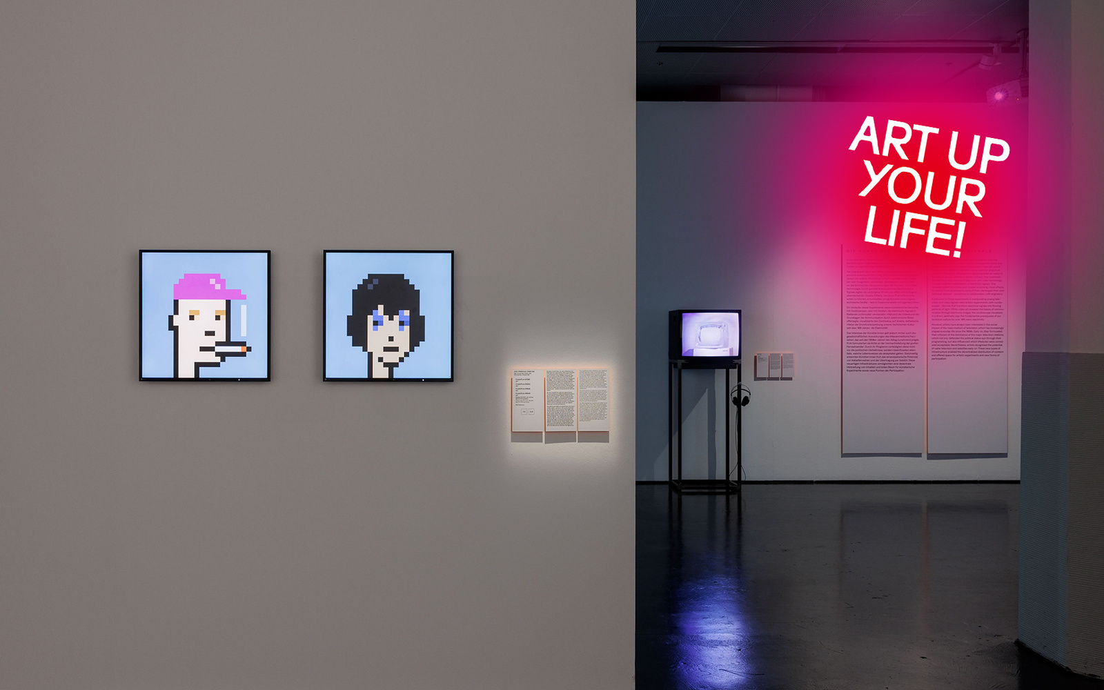 Blick in die Ausstellung mit zwei digitalen Bildern (NFTs) an einer weißen Wand, Infotafeln daneben und einem Monitor mit Kopfhörern im Hintergrund.